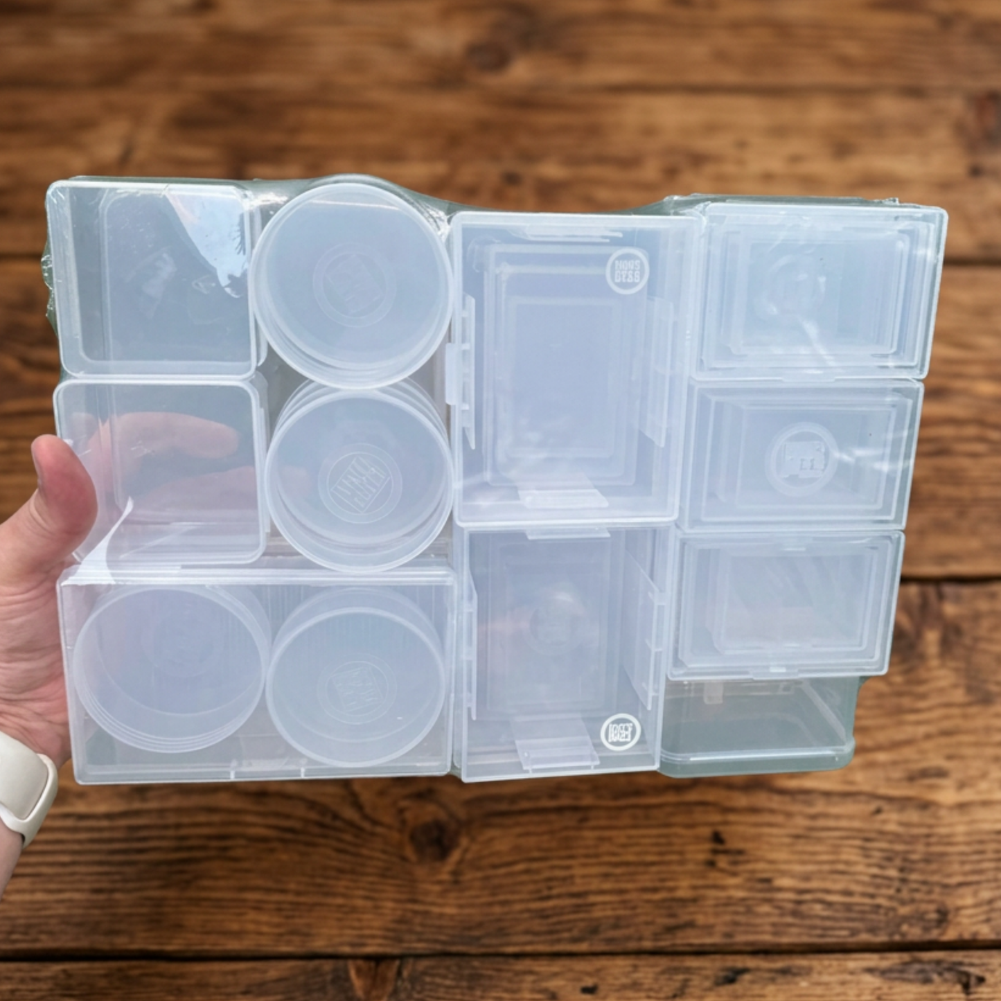 A hand holding a clear plastic organizer tray with multiple small square and round compartments, shown over a rustic wooden surface.